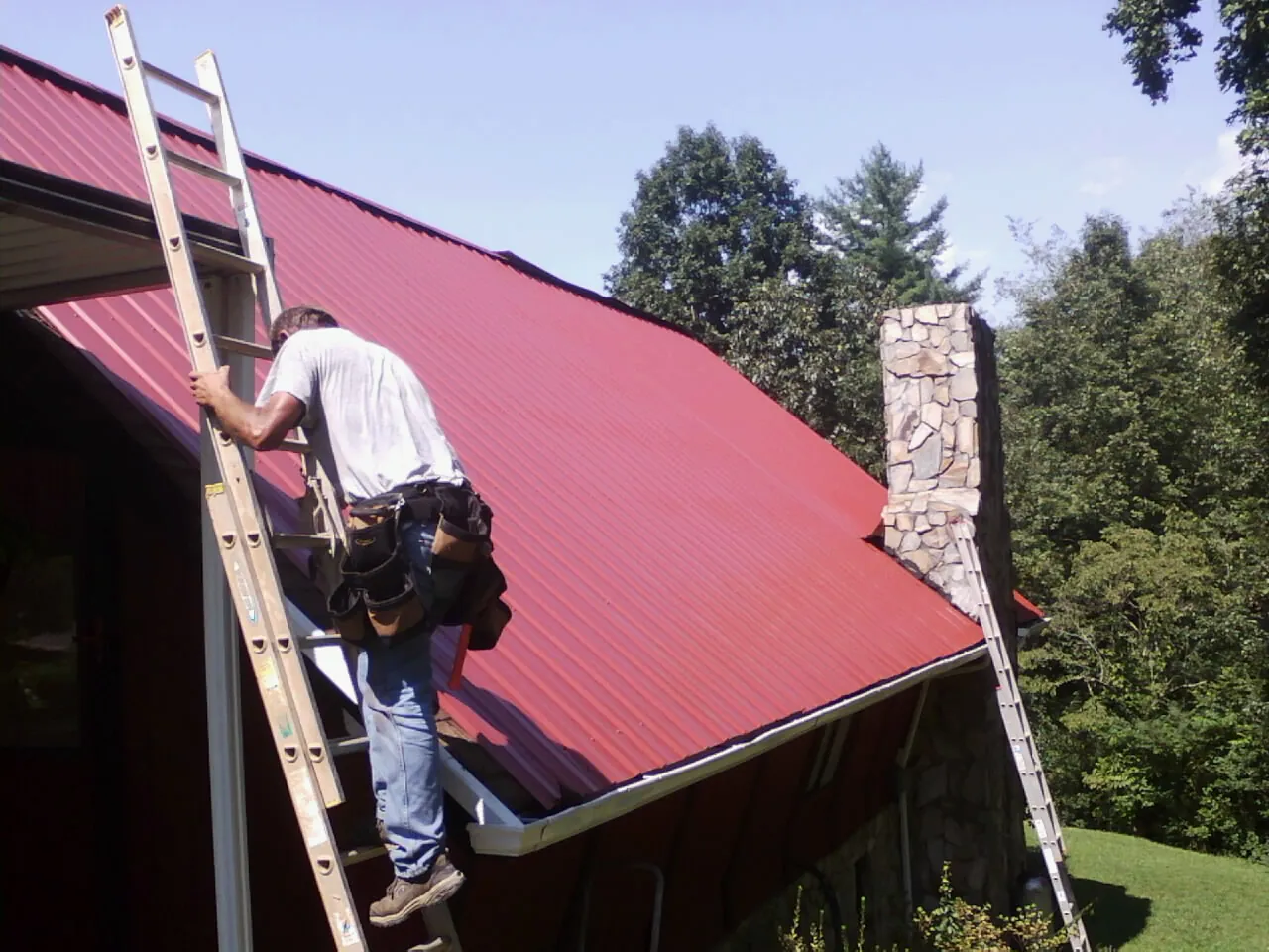 Roofer on a ladder beside a red metal roof and stone chimney — Soffit & Fascia Repair by Roofing Brothers in Butte